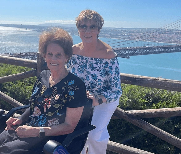 Two women posing on a wooden railing with a coastal city view in the background.
