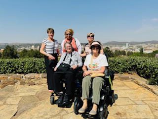 Four women, two in wheelchairs, enjoying a sunny day outdoors with a city view.