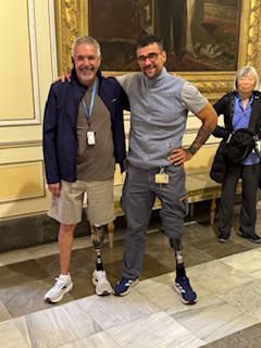 Two men smiling indoors, one casually dressed in shorts, the other in athletic wear with prosthetic legs.