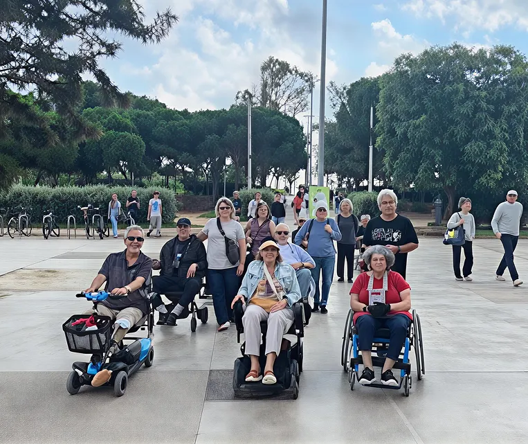Group of people in wheelchairs posing outdoors on a sunny day.