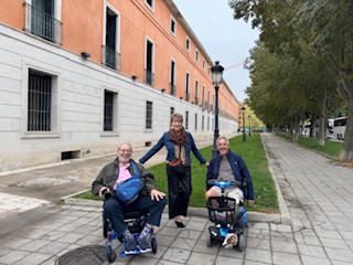 Three elderly people outdoors, two in wheelchairs, with a caregiver standing behind them.