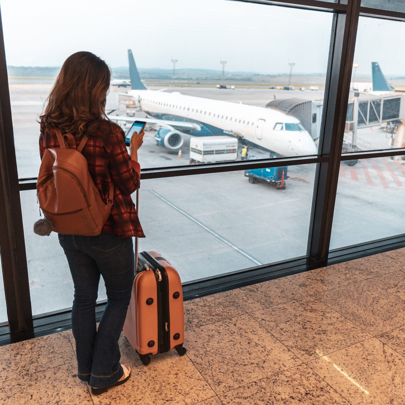 A woman with a suitcase looks at an airplane through the airport window.